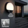 An outdoor wall lamp with a motion sensor is mounted on a textured wall next to a house entrance with stairs. Voir les images du produit
