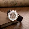 A skeleton watch with a brown leather strap rests on a book on a wooden surface. Náhled obrázku produktu
