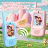 Two children's walkie-talkies, one blue and one pink, are displayed in a whimsical, cartoon-like outdoor setting with rolling hills and a stream. A speech bubble with "hi!" is shown between them. Produktbild-Vorschau
