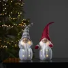 Two illuminated Christmas gnomes stand on a dark surface in front of a blurred Christmas tree with lights. Produktbild-Vorschau