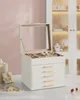 A white jewelry box with a glass lid and gold handles sits on a marble surface, with a pink wall and a display cabinet in the background. Produktbild-Vorschau
