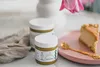 A slice of cheesecake with pistachio topping sits on a pink plate next to two jars of pistachio paste, with a pink tulip in the background. Produktbild-Vorschau