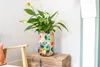 A tall, colorful floral patterned vase with a peace lily plant is placed on a wooden chest next to a chair. Voir les images du produit