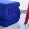 A close-up of a blue bouclé fabric ottoman or bench, folded in half, next to a pink object. Produktbild-Vorschau