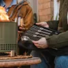 Two people are gathered around a fire pit, with one holding a set of accessories. Produktbild-Vorschau
