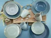 A collection of ceramic dishes and mugs arranged on a light blue surface, with some decorative elements like shells and dried plants. Náhľad obrázku produktu