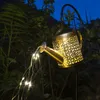 A decorative watering can light in a garden at dusk. Produktbild-Vorschau