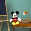 A Mickey Mouse plush toy sits on a wooden desk next to a yellow desk lamp and some pencils, with a chalkboard easel in the background. Voir les images du produit