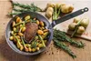 A de Buyer Mineral B frying pan filled with potatoes, asparagus, rosemary, and a piece of duck breast, with additional ingredients and the pan handle visible on a wooden board. Náhľad obrázku produktu