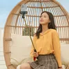 A young woman is sitting in a wicker chair outdoors, holding a selfie stick with a phone attached. Voir les images du produit