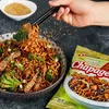 A hand using chopsticks to pick up noodles from a bowl of Chapagetti Jjajang Noodles, with a packet of the product in the foreground. Produktbild-Vorschau