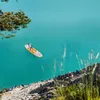 A person paddleboarding on a turquoise lake, with rocky terrain and greenery in the foreground. Náhľad obrázku produktu