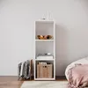 A white, three-shelf cube bookshelf is shown against a plain white wall. The top shelf holds two glass items, one a small glass dome and the other a tall glass bottle. The middle shelf displays a wooden bowl with a textured interior. The bottom shelf conta Produktbild-Vorschau