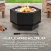 A black hexagonal fire pit with flames burning inside, placed on a patio with wooden chairs and logs nearby. Náhled obrázku produktu