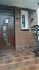 The image shows the entrance to a house, featuring a brown wooden door with decorative glass panels, a small white window, and two black outdoor wall lights mounted on a brown wooden-paneled wall. The ground is tiled with brown and black square tiles, and  Náhľad obrázku produktu