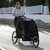 A woman is pushing a black bicycle trailer with two children inside on a paved road. Produktbild-Vorschau
