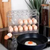 A clear shot of an egg holder filled with eggs, with a frying pan and a fried egg in the foreground. Náhled obrázku produktu