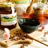 A still life arrangement of honey products, including a jar of honey, a teapot with tea, a bowl of honey, and bee pollen on a wooden board, with flowers. Produktbild-Vorschau