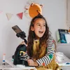 A young girl is using a microscope with a phone attached to it, in a bedroom setting. Produktbild-Vorschau