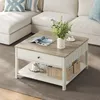 A square coffee table with a drawer and a lower shelf is placed in front of a sectional sofa. The table has a light wood top and a white base. It is decorated with a magazine, a vase of greenery, and a cup of coffee. The lower shelf holds two woven baskets Náhled obrázku produktu