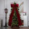 A decorated Christmas tree with a large red bow on top, placed in a room with a fireplace and a lamp. Náhľad obrázku produktu