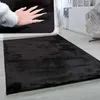 A close-up of a hand touching a black rug, with a wider shot of the rug on a floor in the background. Produktbild-Vorschau