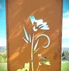 A decorative metal screen with a floral and butterfly cutout design, set against a natural outdoor background with trees and buildings. Produktbild-Vorschau