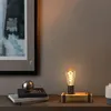A vintage-style desk lamp with a wooden base and exposed bulb sits on a shelf next to a stack of books and a metallic sphere. Produktbild-Vorschau