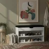 A shoe cabinet with a padded seat, placed in a hallway or entryway, next to a plant and a coat rack. Produktbild-Vorschau