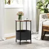 A small round side table with a wooden top and a grey fabric basket underneath, placed next to a chair and a plant in a bright room. Náhled obrázku produktu
