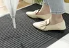 A person's feet wearing beige shoes standing on a grey ribbed doormat. Anteprima dell’immagine prodotto