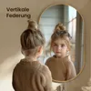 A young girl looks into an oval mirror, her reflection showing her face and upper body. The mirror has a gold frame and is mounted on a wall. Náhled obrázku produktu