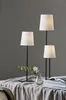 Three table lamps of varying heights are arranged on a table covered with a textured cloth. A small vase with greenery and a book are also present. Podgląd zdjęcia produktu