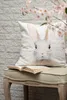 A decorative pillow with a bunny print sits on a bench next to a book, with a branch of pink blossoms in the foreground. Podgląd zdjęcia produktu
