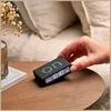 A hand is shown adjusting a black digital alarm clock on a wooden table next to a book and a vase of dried flowers. Produktbild-Vorschau