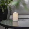 A white LED candle sits on a glass-topped table next to a potted plant. Produktbild-Vorschau