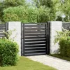 A black metal garden gate is shown in an outdoor setting, with a paved walkway leading to it, surrounded by greenery and stone pillars. Produktbild-Vorschau