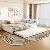 A bedroom scene featuring a beige upholstered daybed with a pull-out trundle bed, decorated with pillows and blankets. The bed has integrated LED lighting and a bookshelf unit behind it. Náhled obrázku produktu