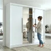 A woman is opening a sliding door of a white wardrobe with mirrored panels. The wardrobe interior is visible, showing clothes on hangers and shelves. The room appears to be a bedroom. Produktbild-Vorschau