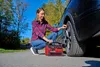A woman is using a portable air compressor to inflate a car tire outdoors. Produktbild-Vorschau