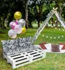 An outdoor picnic setup with a pallet sofa, cushions, balloons, and a teepee. Produktbild-Vorschau