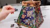 A hand is shown placing a puzzle piece onto a partially assembled 3D wooden puzzle shaped like a pyramid with colorful, intricate designs. Several loose puzzle pieces are scattered around the base of the puzzle. Náhľad obrázku produktu