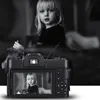 A black digital camera is shown in the foreground, displaying a picture of a young girl on its screen. The background is a black and white image of the same girl, appearing as if she is being photographed. Podgląd zdjęcia produktu