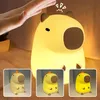 A hand is shown interacting with a glowing capybara-shaped night light. Smaller inset images show the product in different lighting conditions. Podgląd zdjęcia produktu