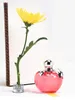 A yellow flower in a small metal holder next to a pink apple-shaped perfume bottle. Anteprima dell’immagine prodotto