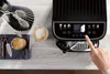 A hand is shown pressing a button on a black coffee machine, with a cup of coffee and a cookie nearby. Voir les images du produit