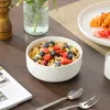 A bowl of cereal with strawberries and blueberries is placed on a wooden table next to a newspaper and cutlery. A glass vase with flowers is in the background. Produktbild-Vorschau