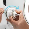 Hands holding a flexible LED magnifying mirror with a suction cup, demonstrating its attachment to a reflective surface. Podgląd zdjęcia produktu