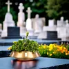 A decorative bowl filled with plants sits on a dark surface in a cemetery, with gravestones and flowers in the background. Produktbild-Vorschau