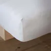 A close-up view of a white fitted sheet, showing the corner seam and texture of the fabric. The sheet is draped over what appears to be a bed frame or mattress base, with wooden floorboards visible below. Náhľad obrázku produktu